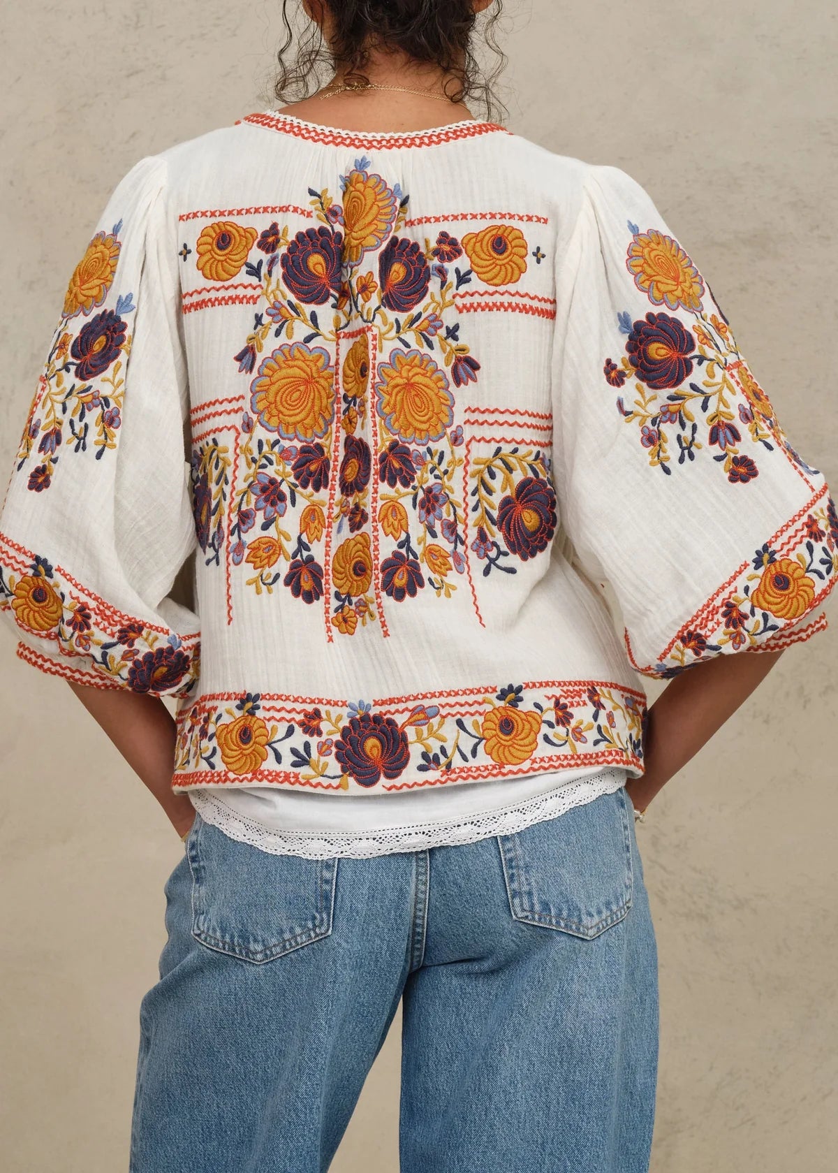 Person wearing a white blouse with colorful floral embroidery and blue jeans against a beige background