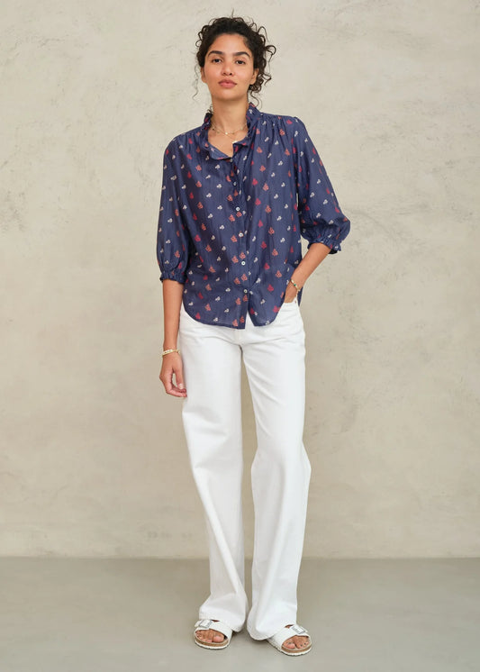 Woman wearing a blue patterned blouse and white pants against a plain background