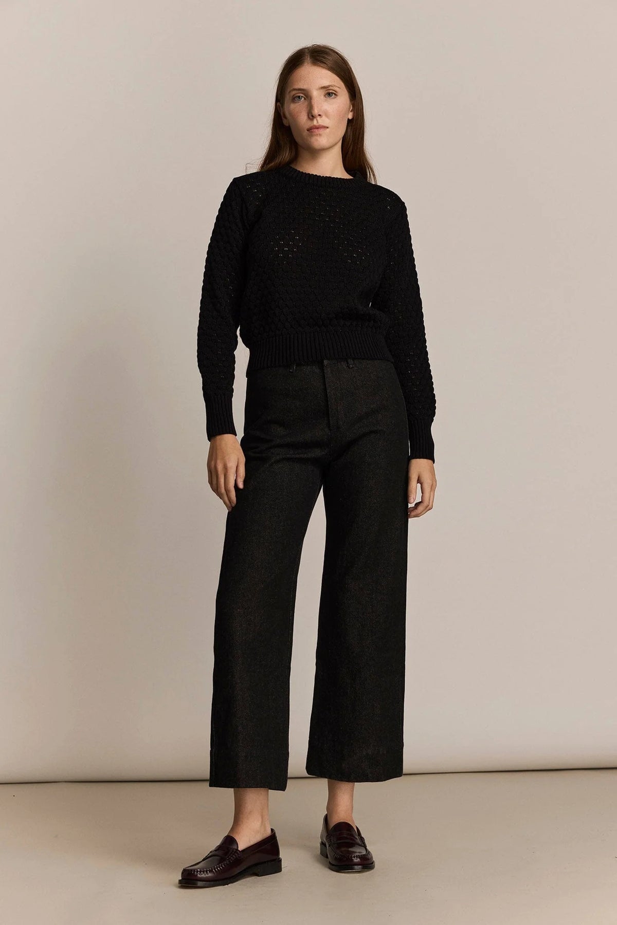A woman stands against a plain beige studio background wearing a black textured knit sweater, black cropped wide-leg trousers, and dark brown loafers.