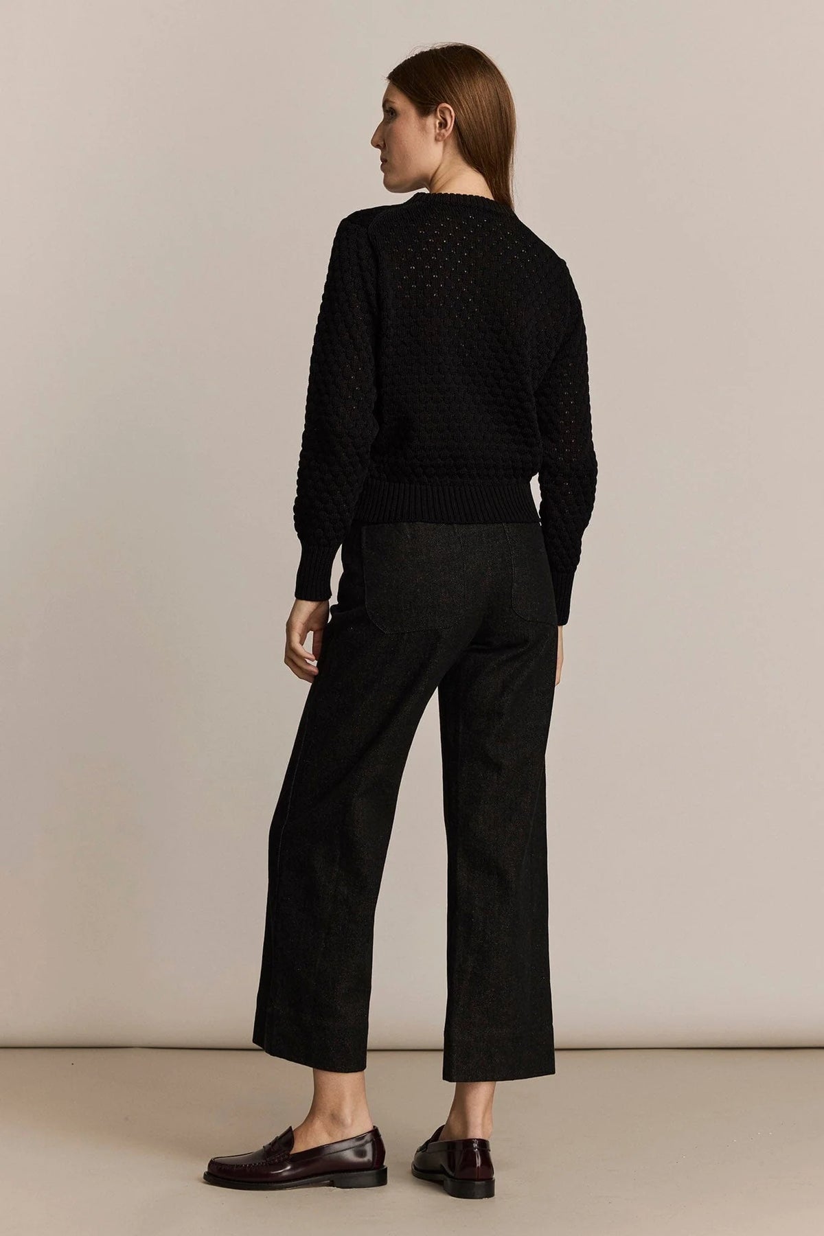 A woman is shown from the back wearing a black textured knit sweater and matching black wide-leg cropped trousers, paired with dark brown leather loafers, against a plain beige studio background.