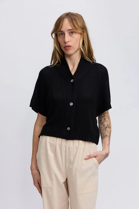 A woman modeling a short-sleeve black knit button-front cardigan with a shawl collar, paired with cream-colored trousers, against a plain light gray studio background.