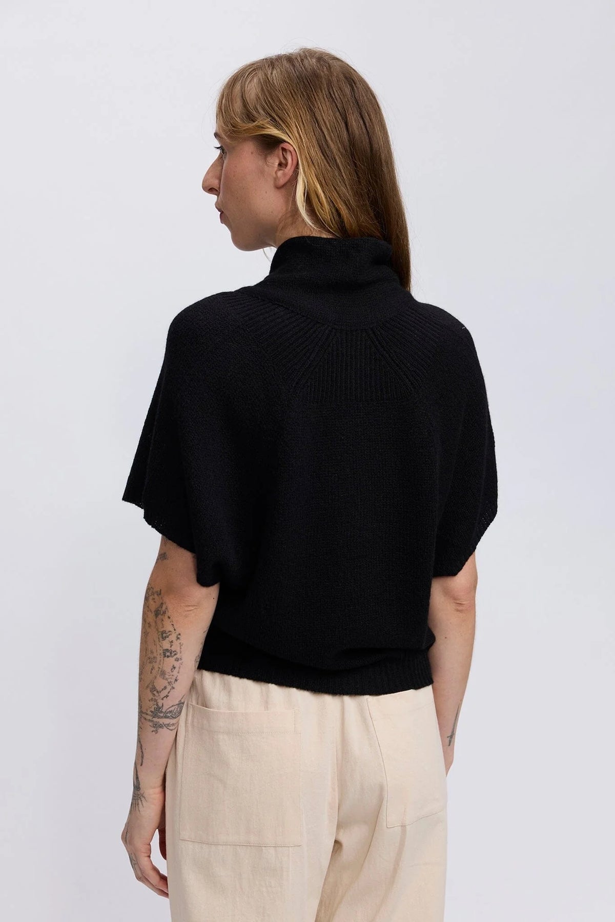 A woman shown from the back wearing a black short-sleeve ribbed mock-neck knit top and light beige trousers, photographed against a plain light gray studio background.
