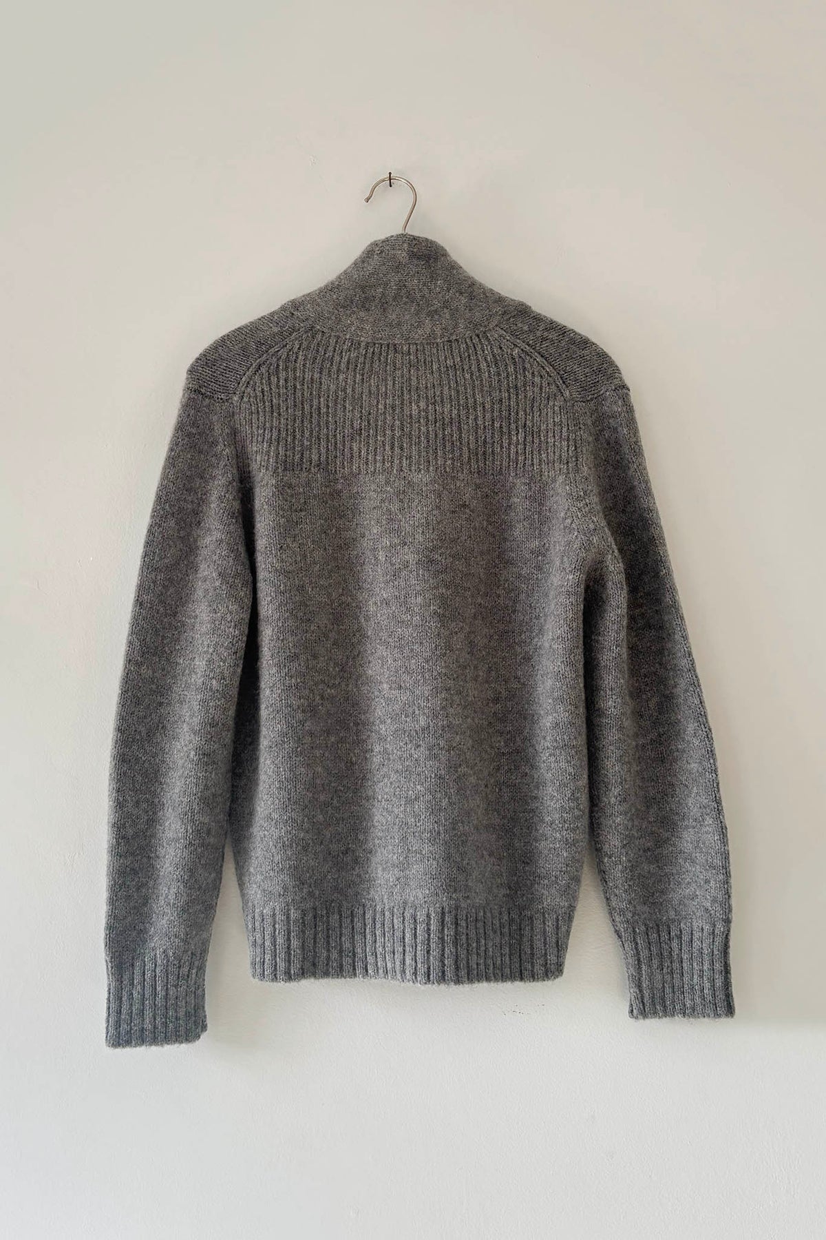 A gray knit sweater with a ribbed mock neck, shoulders, cuffs, and hem, hanging on a metal hanger against a plain off-white wall.