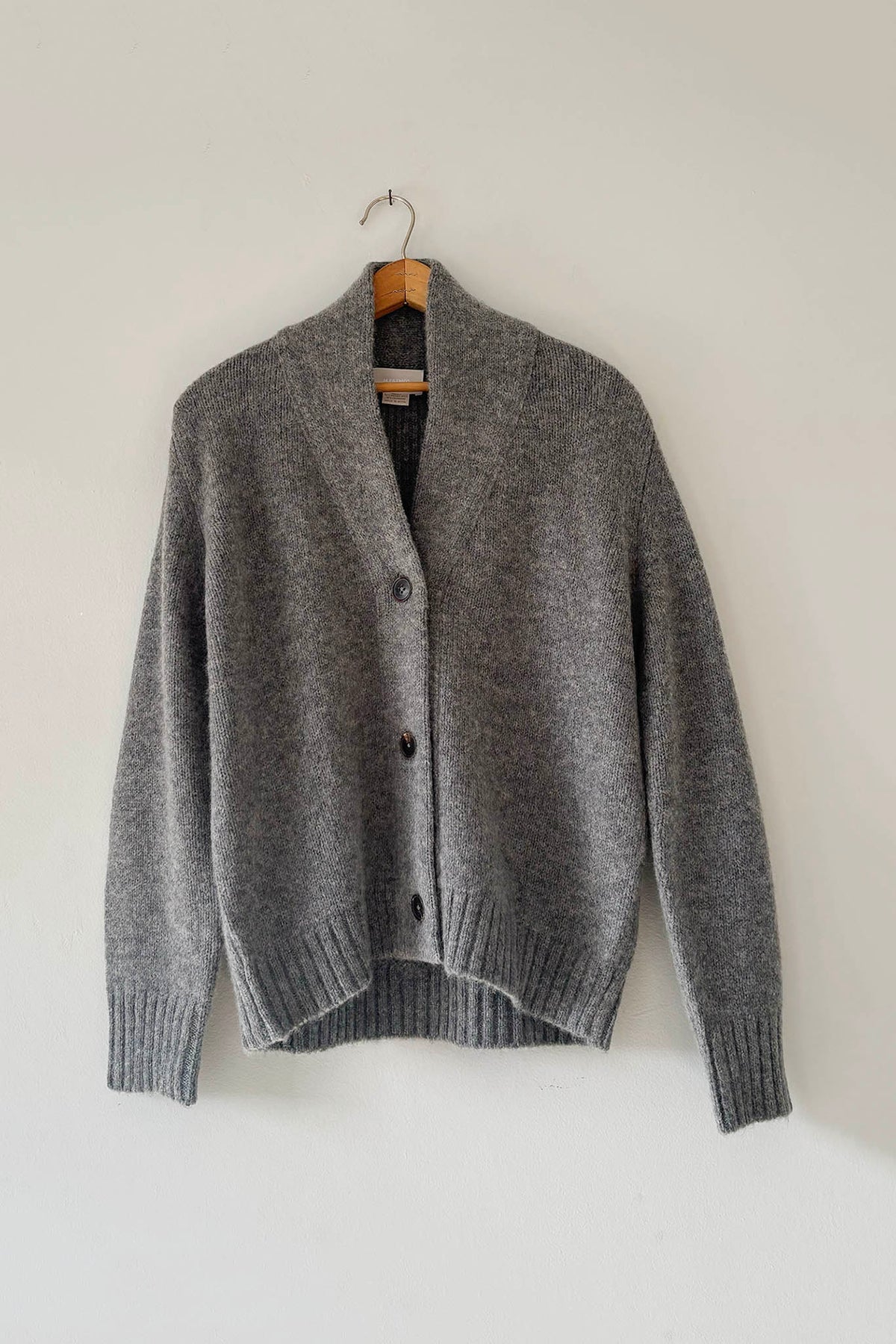 A gray knit cardigan sweater hanging on a wooden hanger against a plain off-white wall.