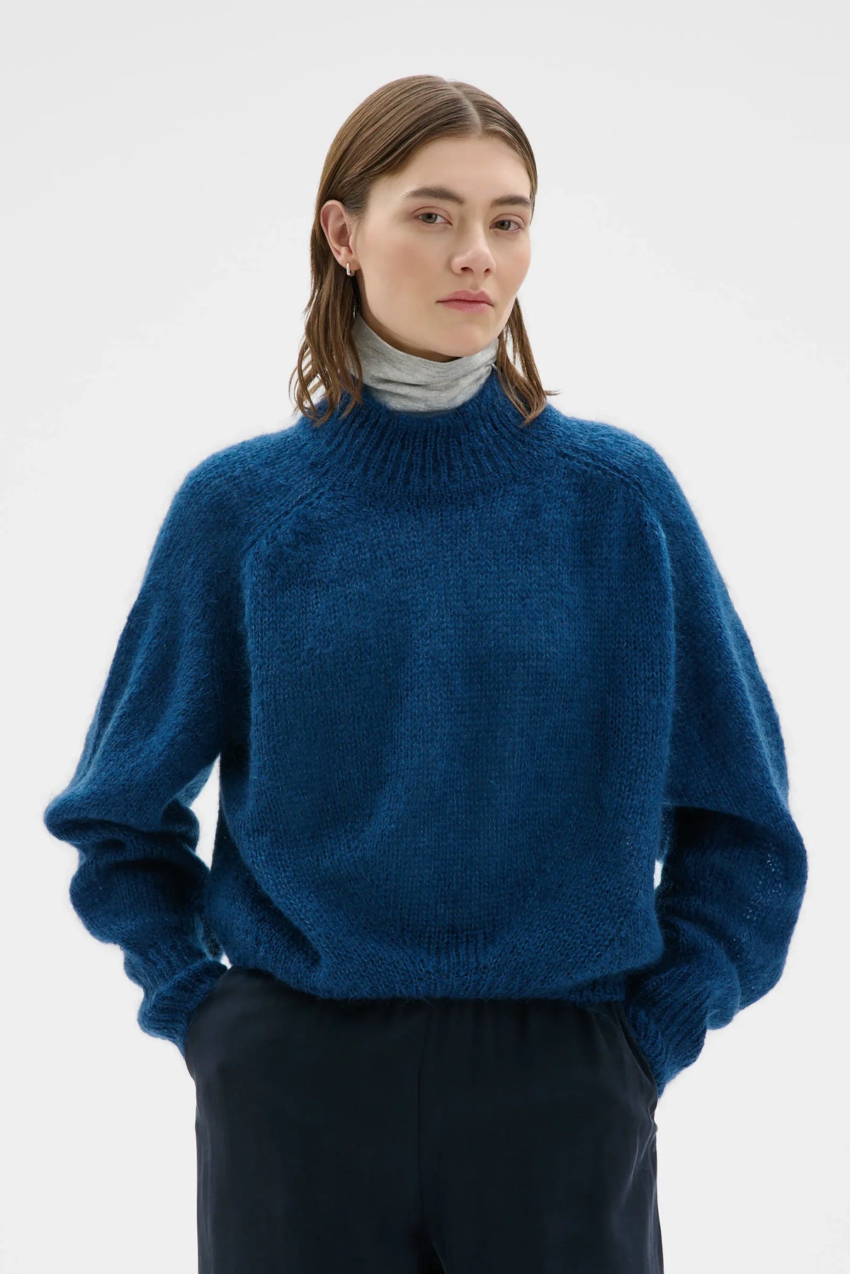 A woman wearing a textured, chunky knit blue turtleneck sweater over a light gray turtleneck, paired with dark trousers, against a plain light gray studio background.