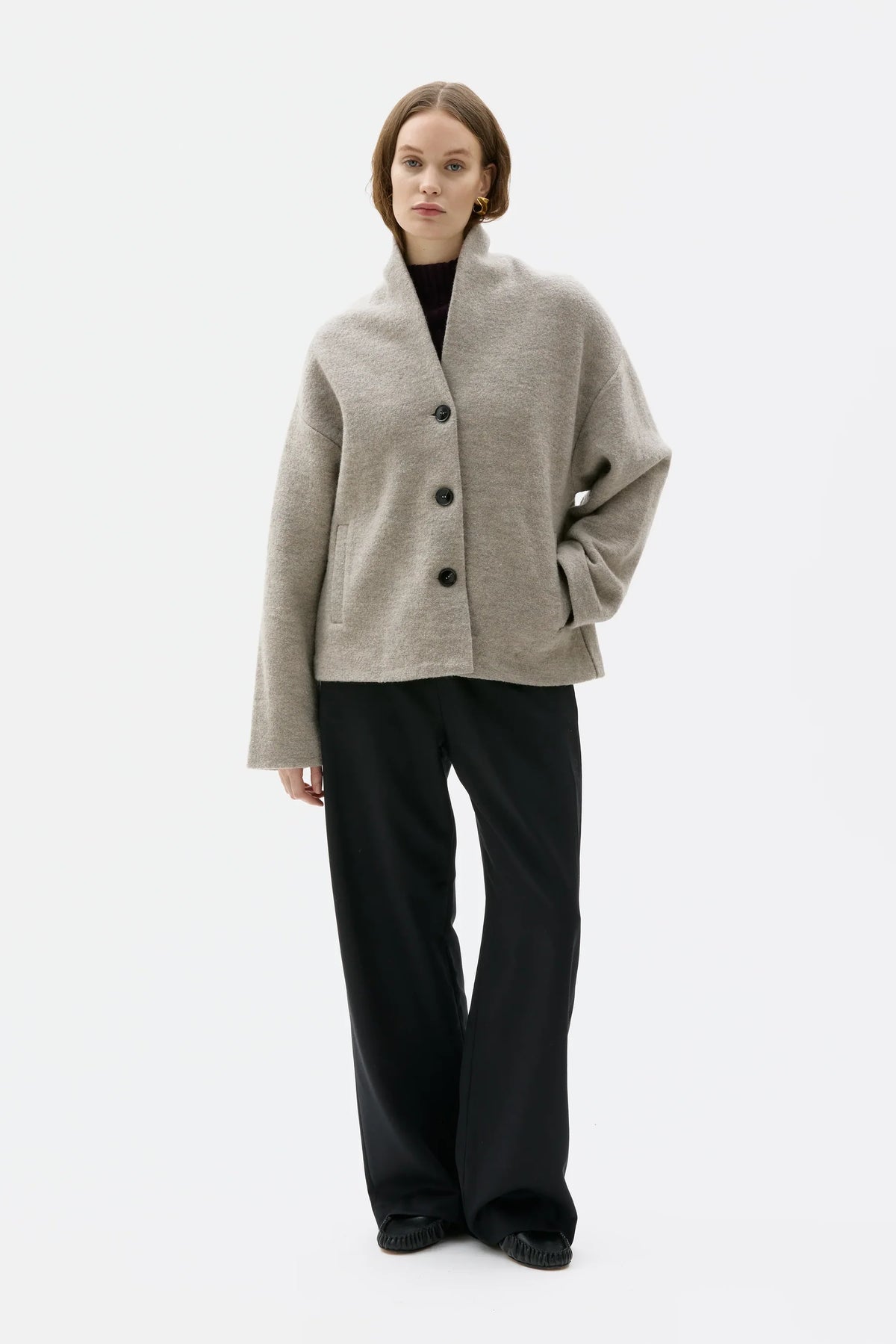 A model stands against a plain white studio background wearing a light beige open-front cardigan with dark buttons, black wide-leg trousers, and black shoes.