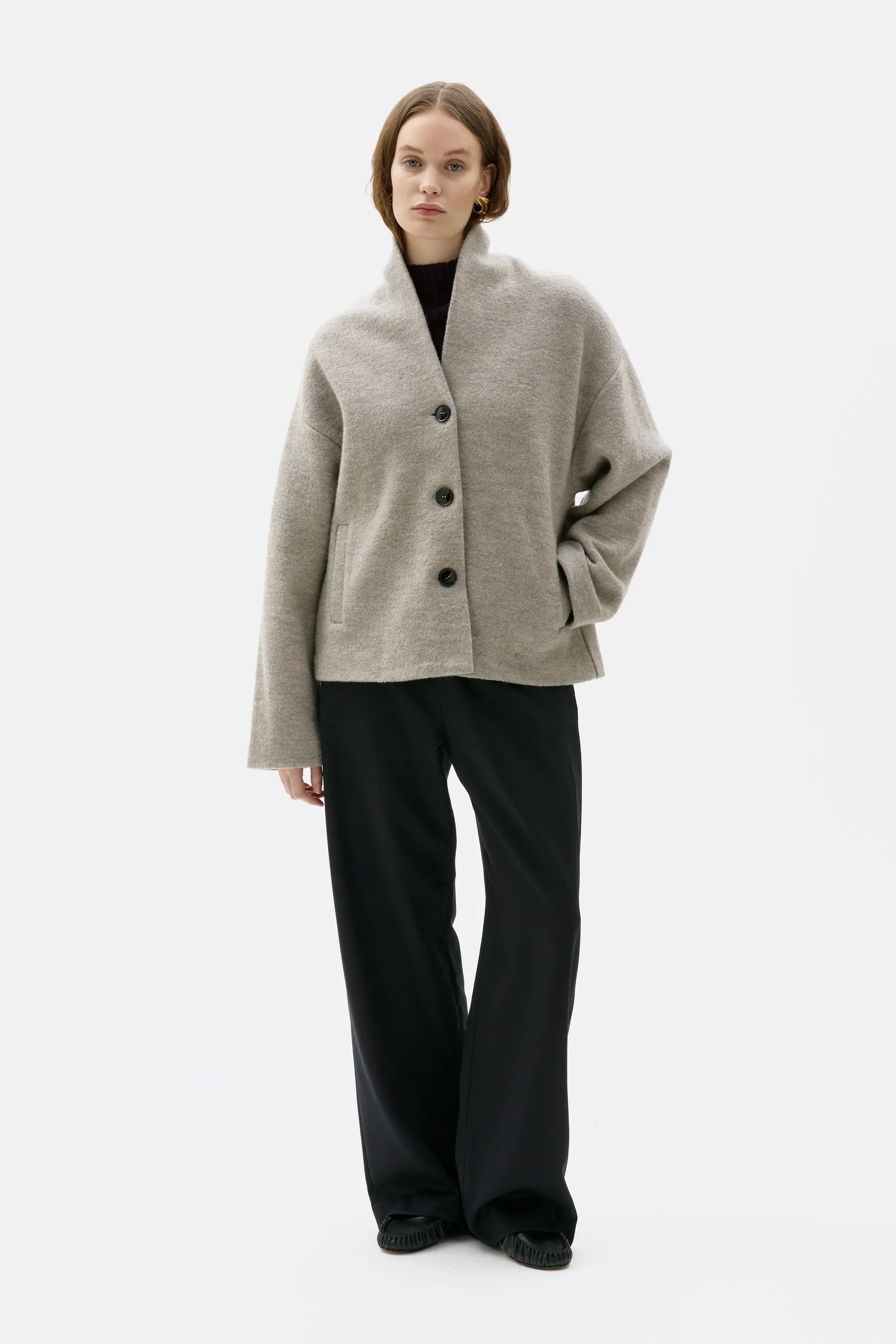 A model stands against a plain white studio background wearing a light beige open-front cardigan with dark buttons, black wide-leg trousers, and black shoes.