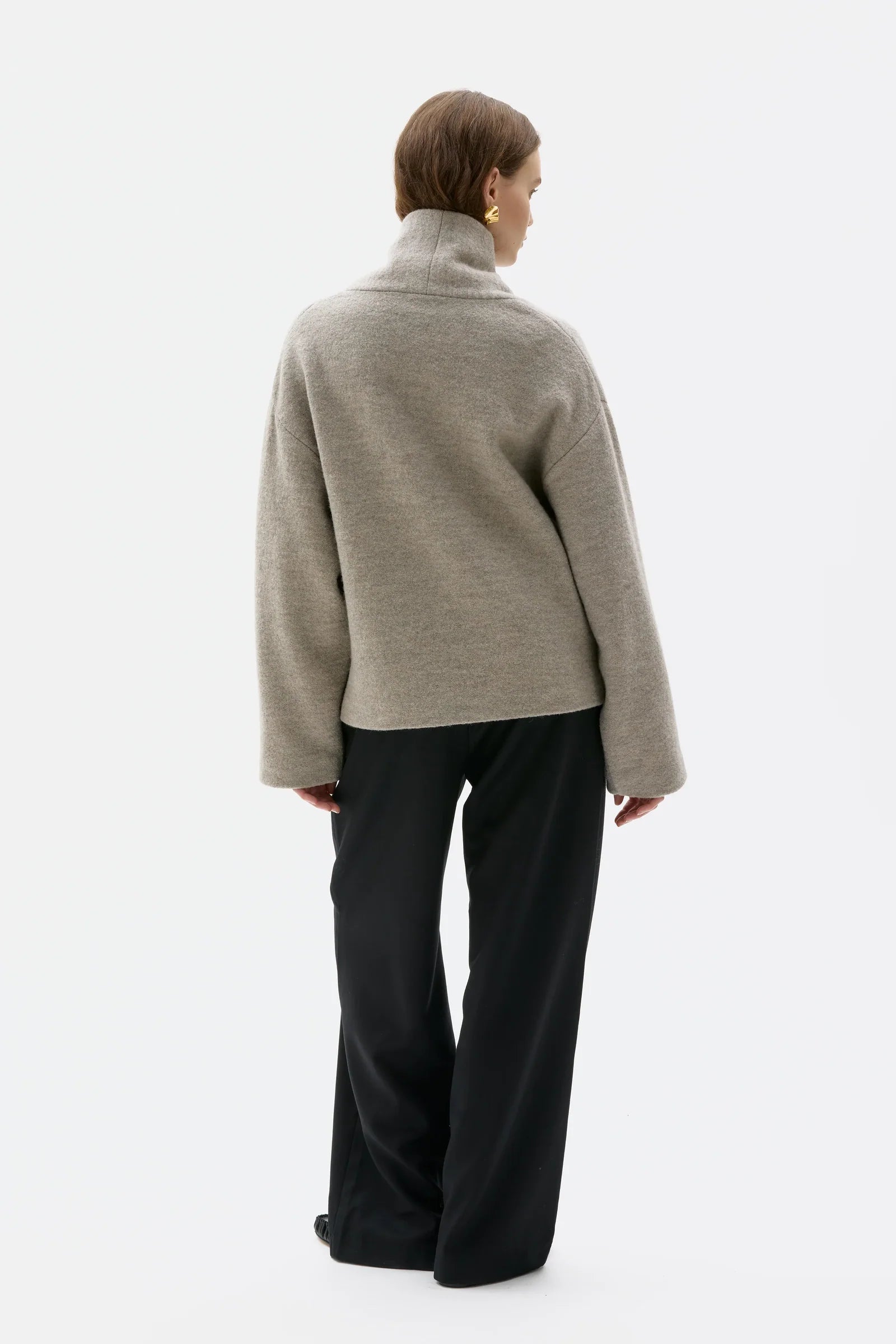 A model shown from the back wearing a light gray turtleneck sweater and black wide-leg trousers against a plain white studio background.