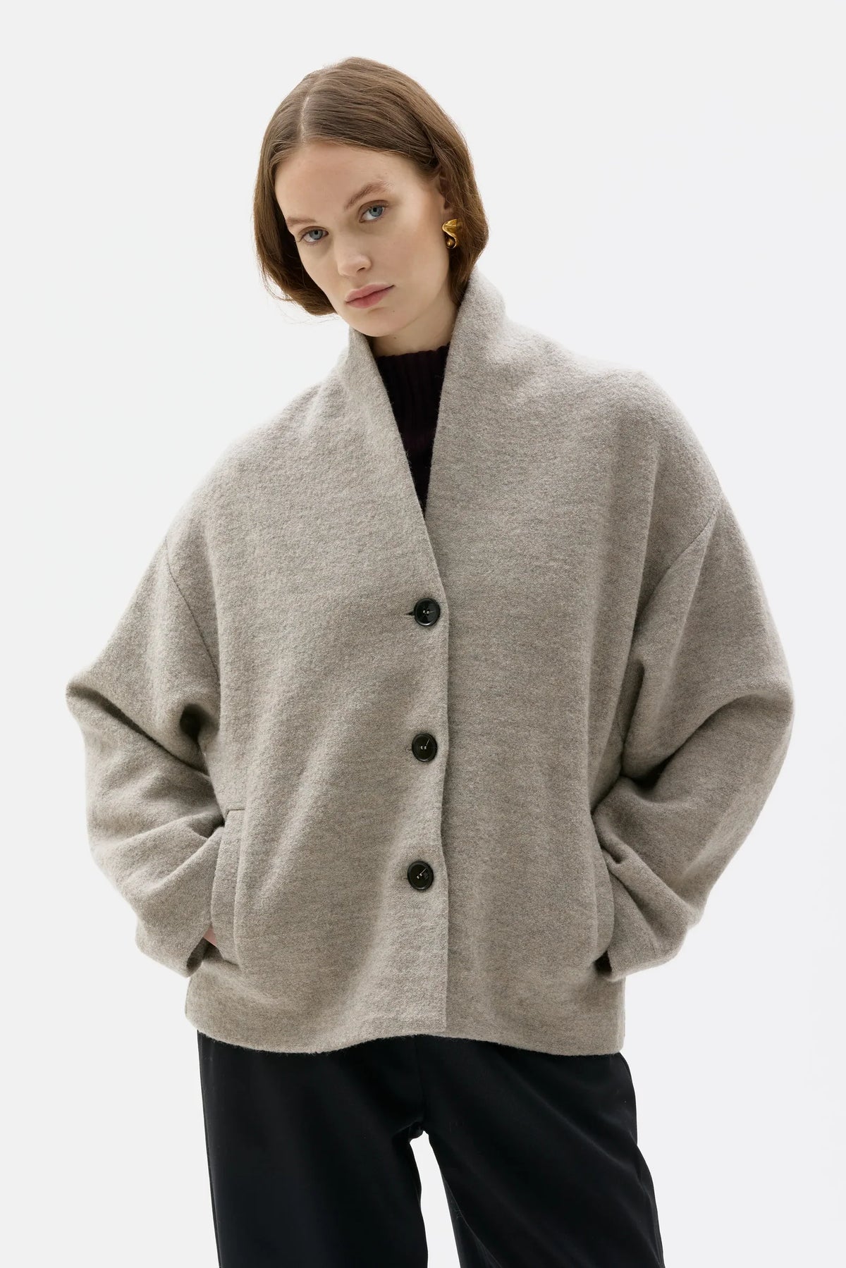 A woman wearing a light gray oversized knit cardigan with a shawl collar and dark buttons, paired with black pants, against a plain light gray studio background.
