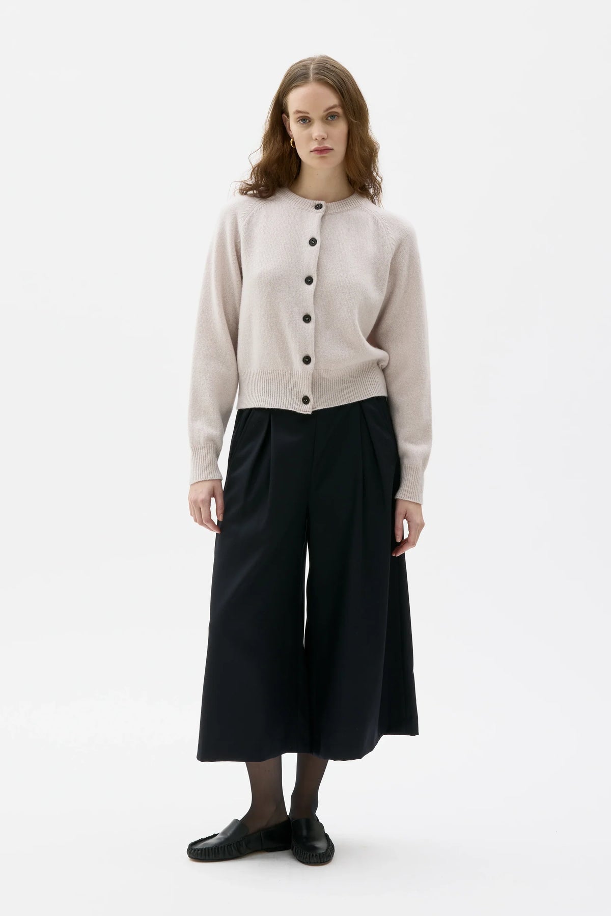 Woman wearing a beige cardigan, black pants, and black shoes on a white background