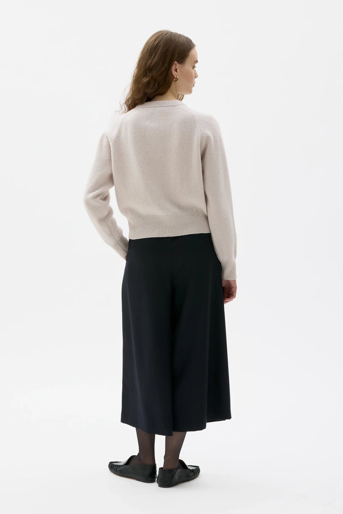 A woman shown from the back wearing a light beige knit sweater and a long black skirt, standing against a plain white studio background.