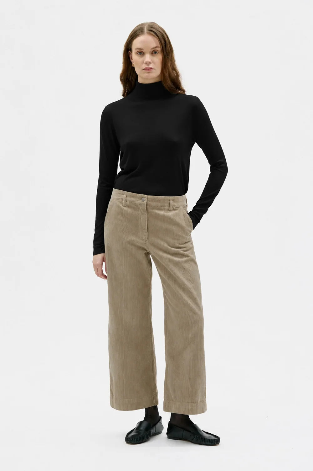 A woman modeling a black long-sleeve turtleneck top and beige wide-leg cropped trousers, paired with black shoes, against a plain white studio background.
