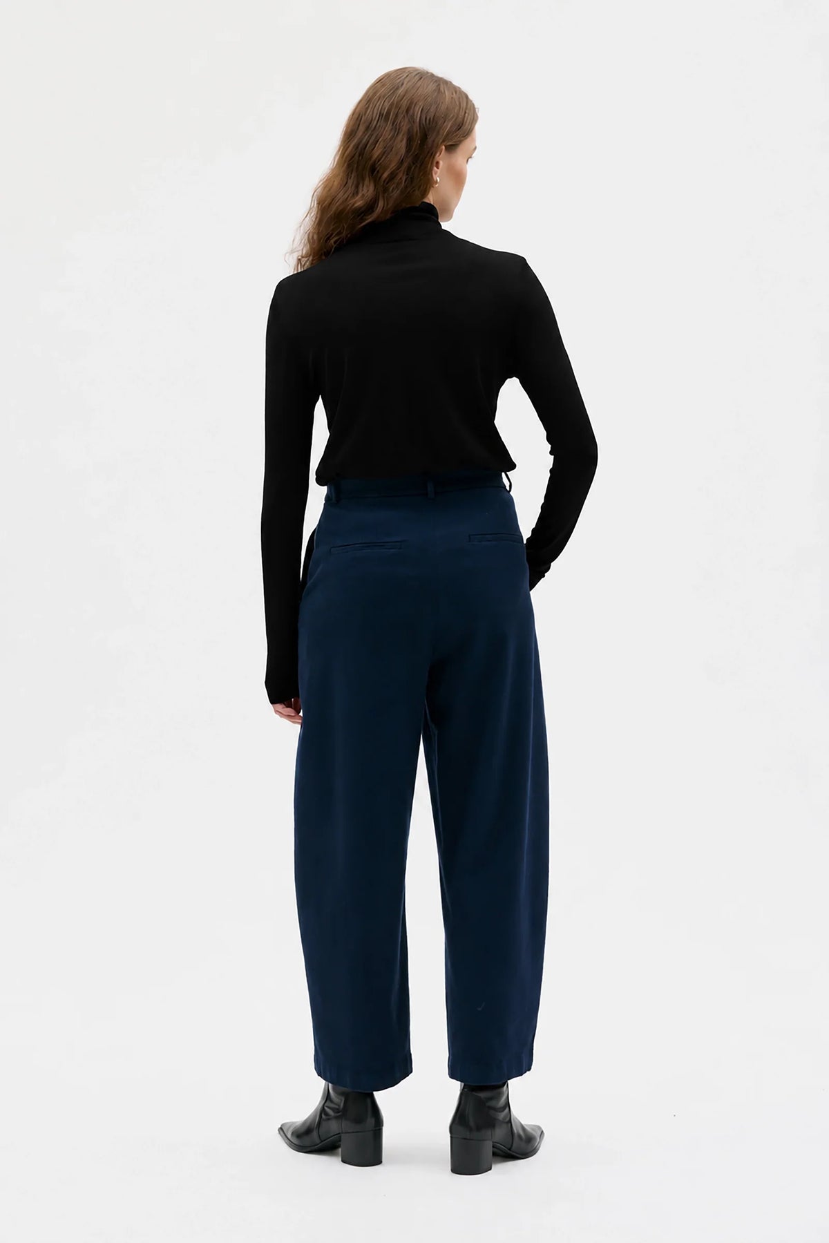 A woman is shown from the back wearing a black long-sleeve mock neck top and navy blue wide-leg trousers, paired with black ankle boots, against a plain white studio background.