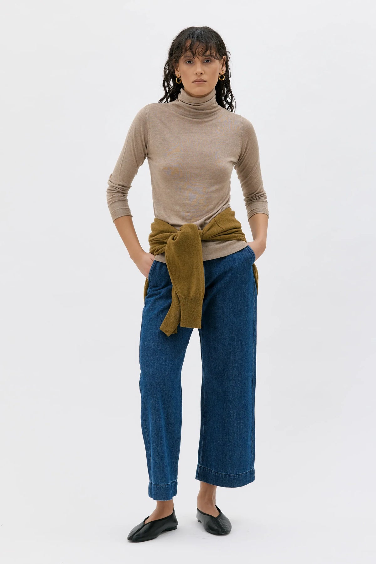 A model stands against a plain white studio background wearing a beige ribbed turtleneck top, wide-leg blue jeans, and black flats, with an olive green sweater tied around her waist.