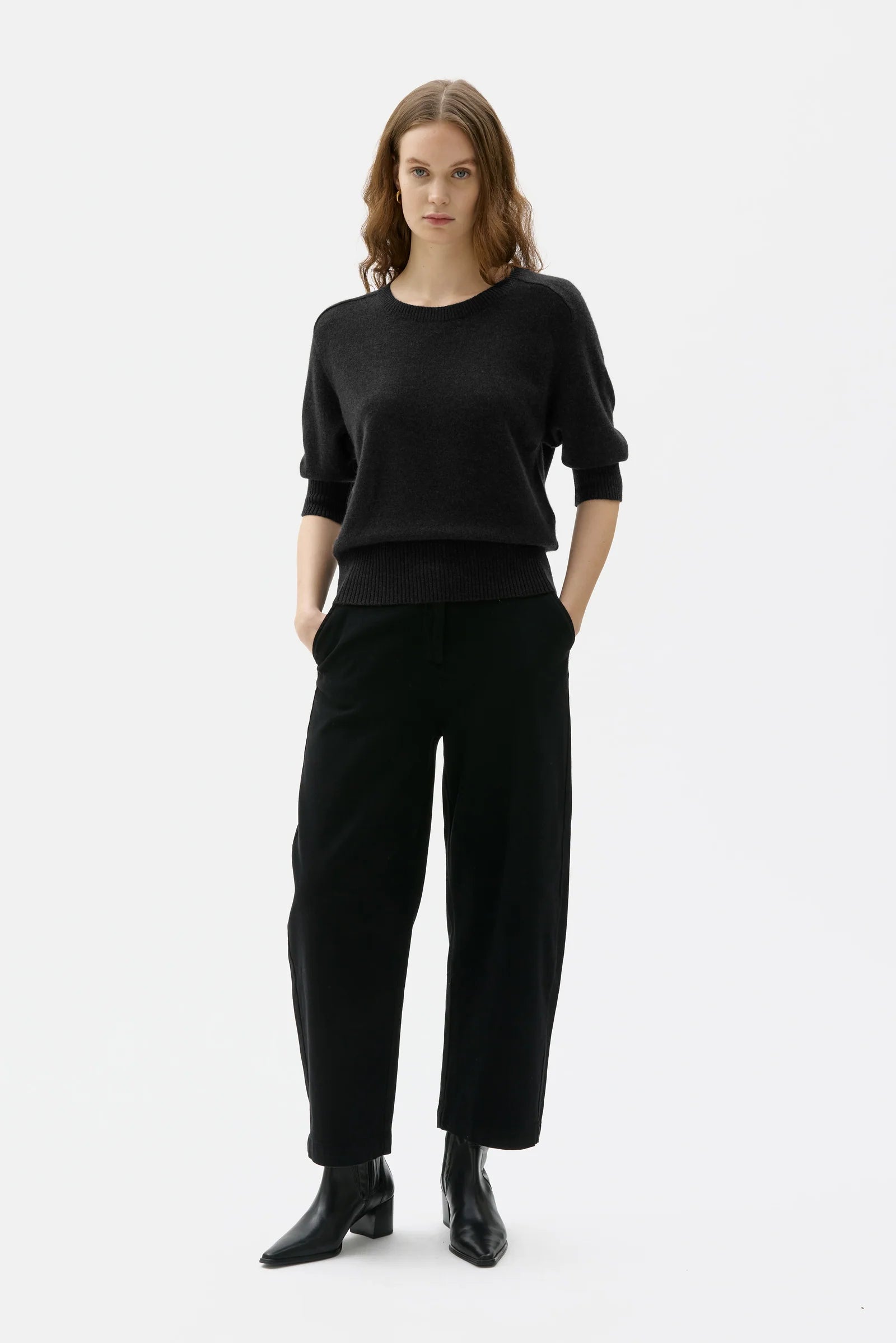 A woman modeling a black long-sleeve knit sweater and matching black wide-leg trousers, paired with black ankle boots, against a plain light gray studio background.