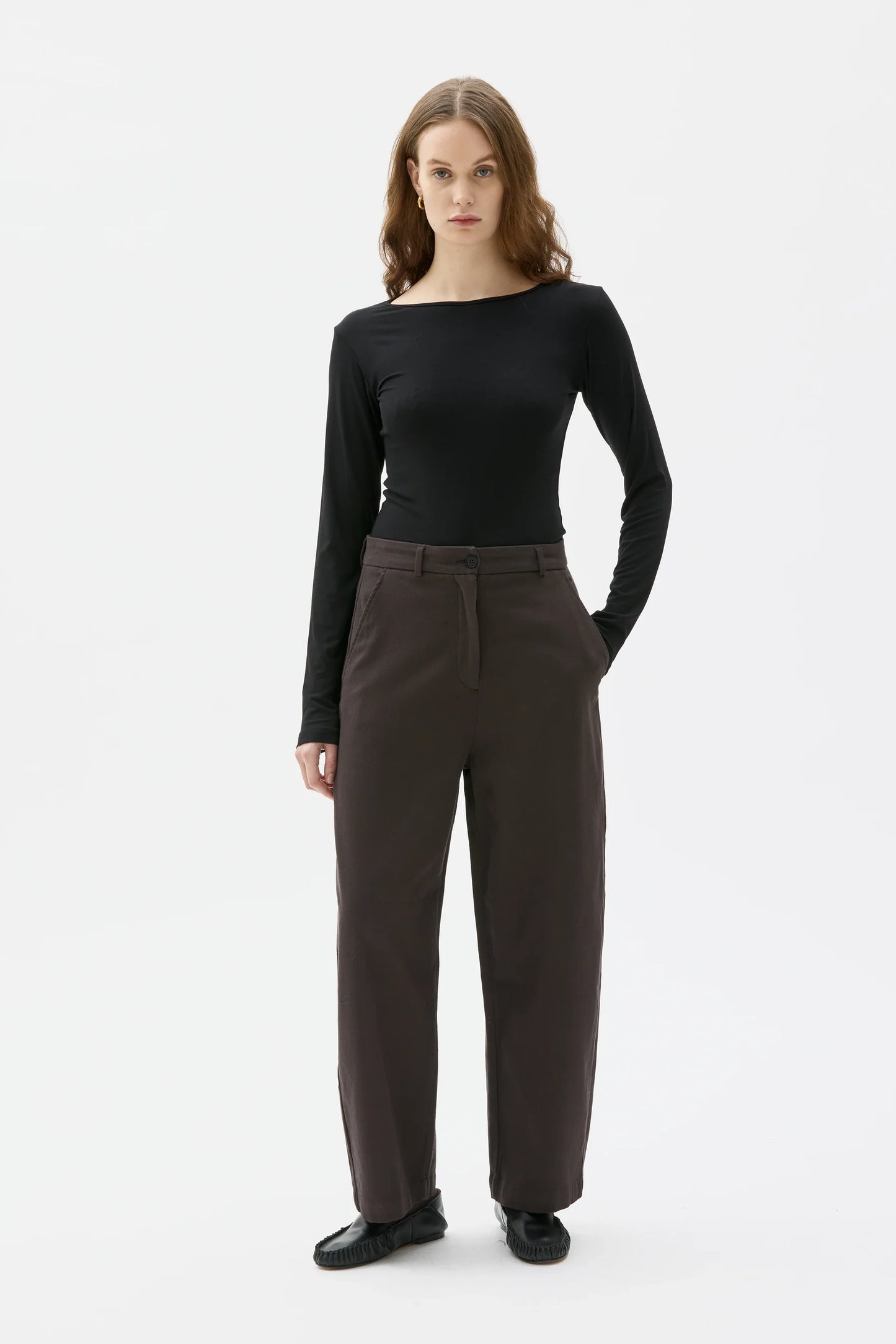 A woman modeling a black long-sleeve boat-neck top and dark brown wide-leg trousers against a plain white studio background.