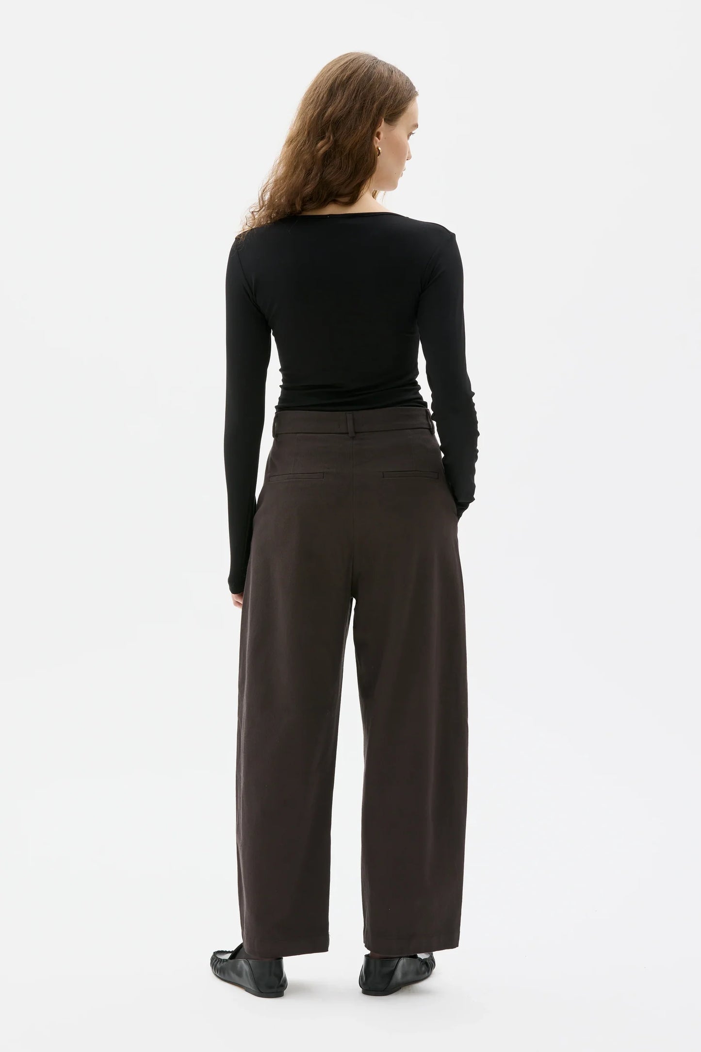 A woman shown from the back wearing a black long-sleeve top and dark brown wide-leg trousers against a plain white studio background.