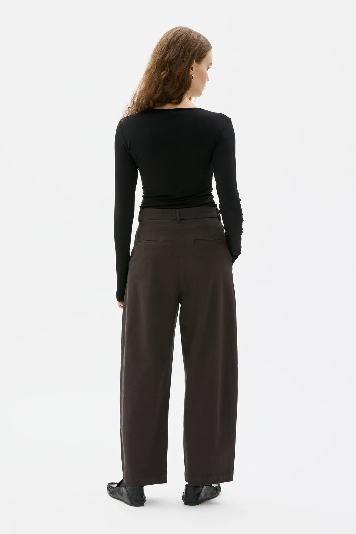 A woman shown from the back wearing a black long-sleeve top and dark brown wide-leg trousers against a plain white studio background.