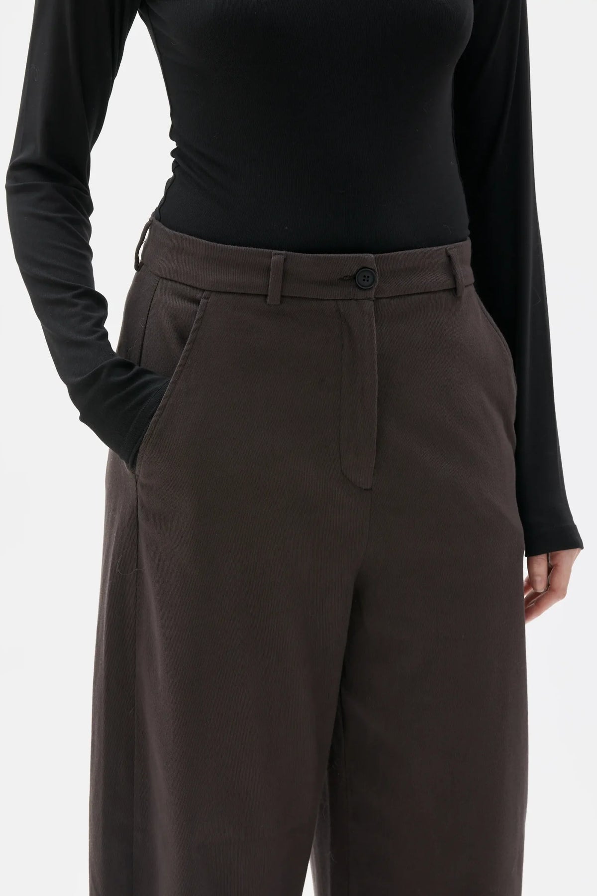 A close-up of a model wearing dark brown wide-leg trousers with a button waistband and belt loops, paired with a black long-sleeve top against a light gray background.