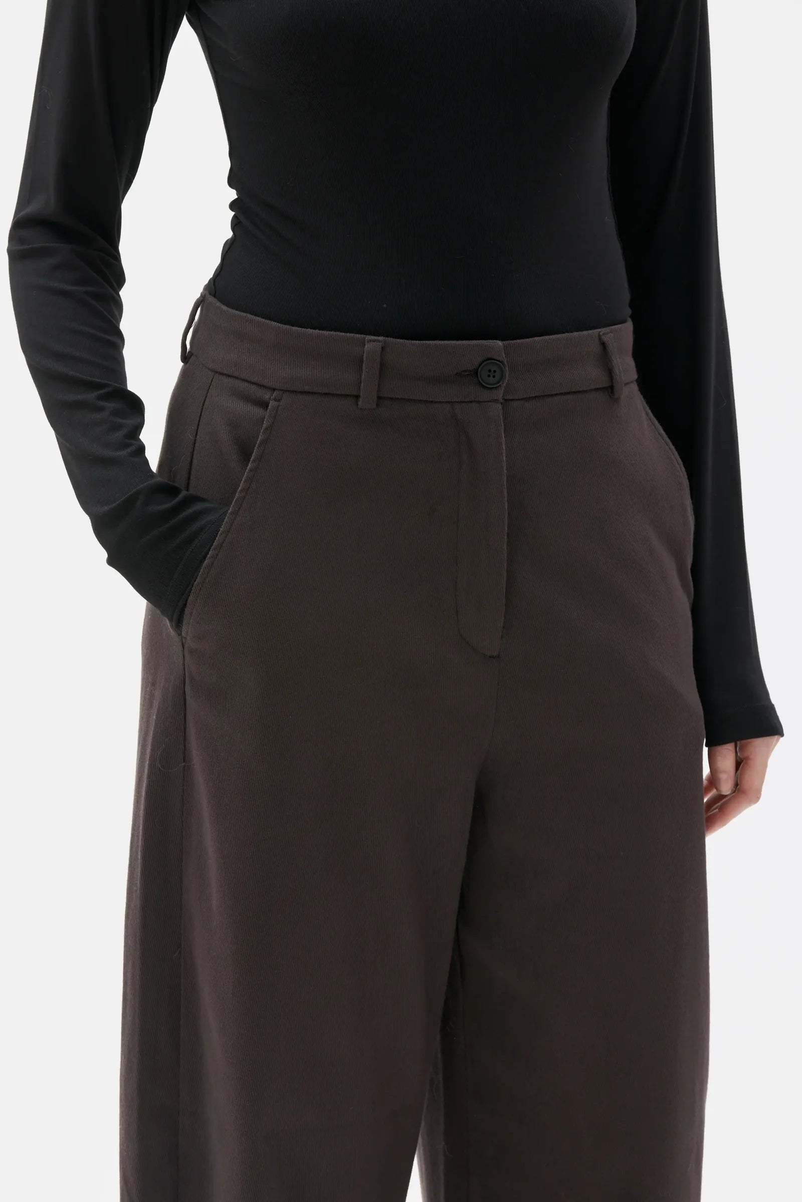 A close-up of a model wearing dark brown wide-leg trousers with a button waistband and belt loops, paired with a black long-sleeve top against a light gray background.