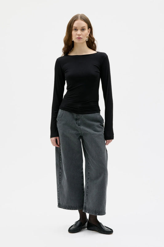 A woman modeling a black long-sleeve boat-neck top with wide-leg charcoal gray jeans and black loafers, standing against a plain white studio background.