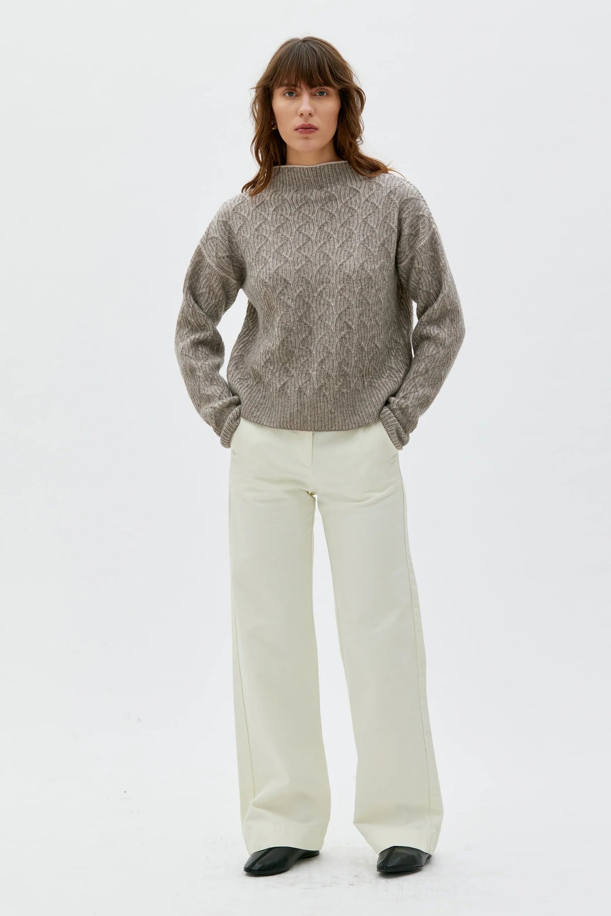 A woman stands against a plain white studio background wearing a textured taupe-gray cable-knit sweater and off-white wide-leg trousers.