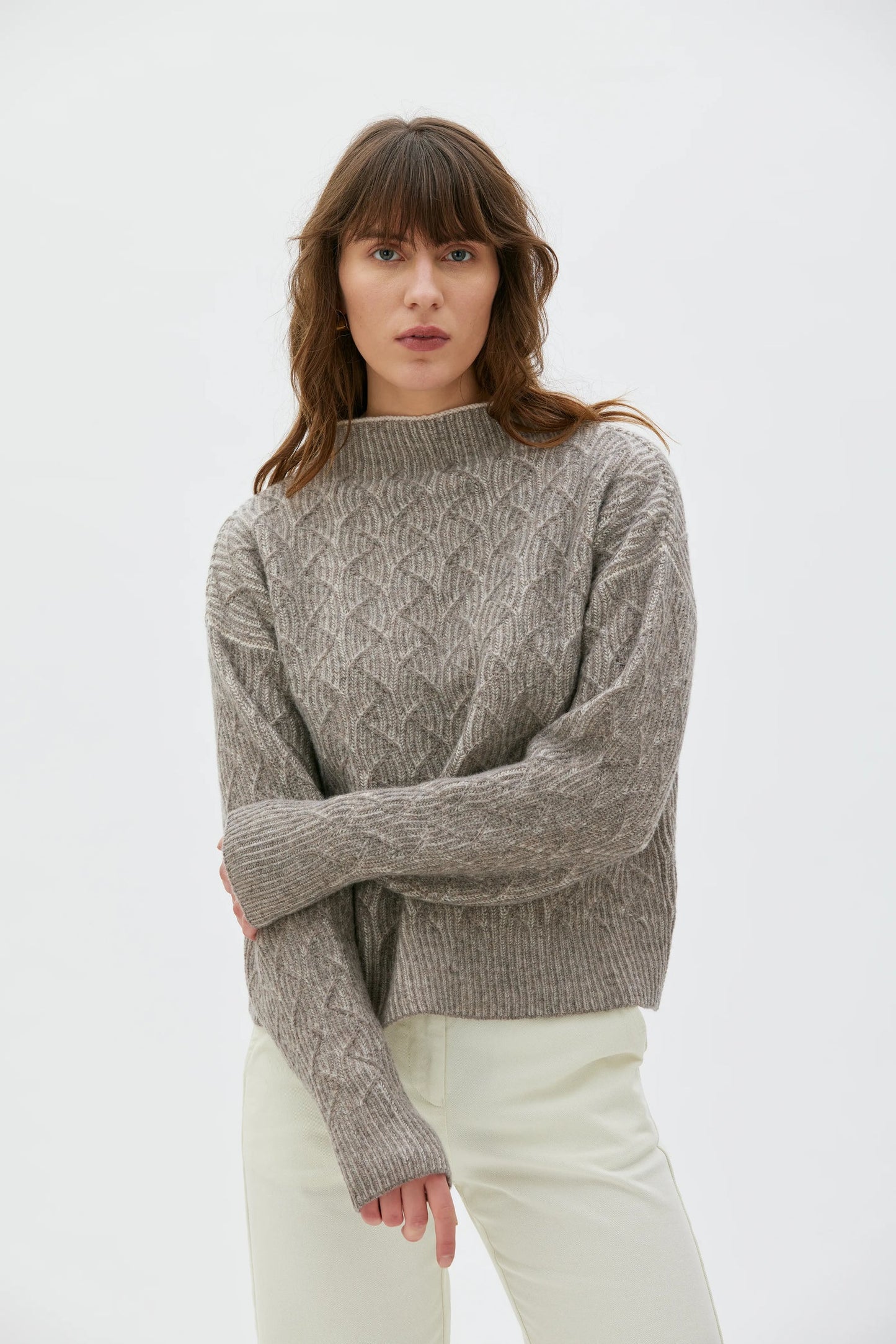 A woman wearing a taupe cable-knit mock-neck sweater and cream trousers, standing against a plain white studio background.