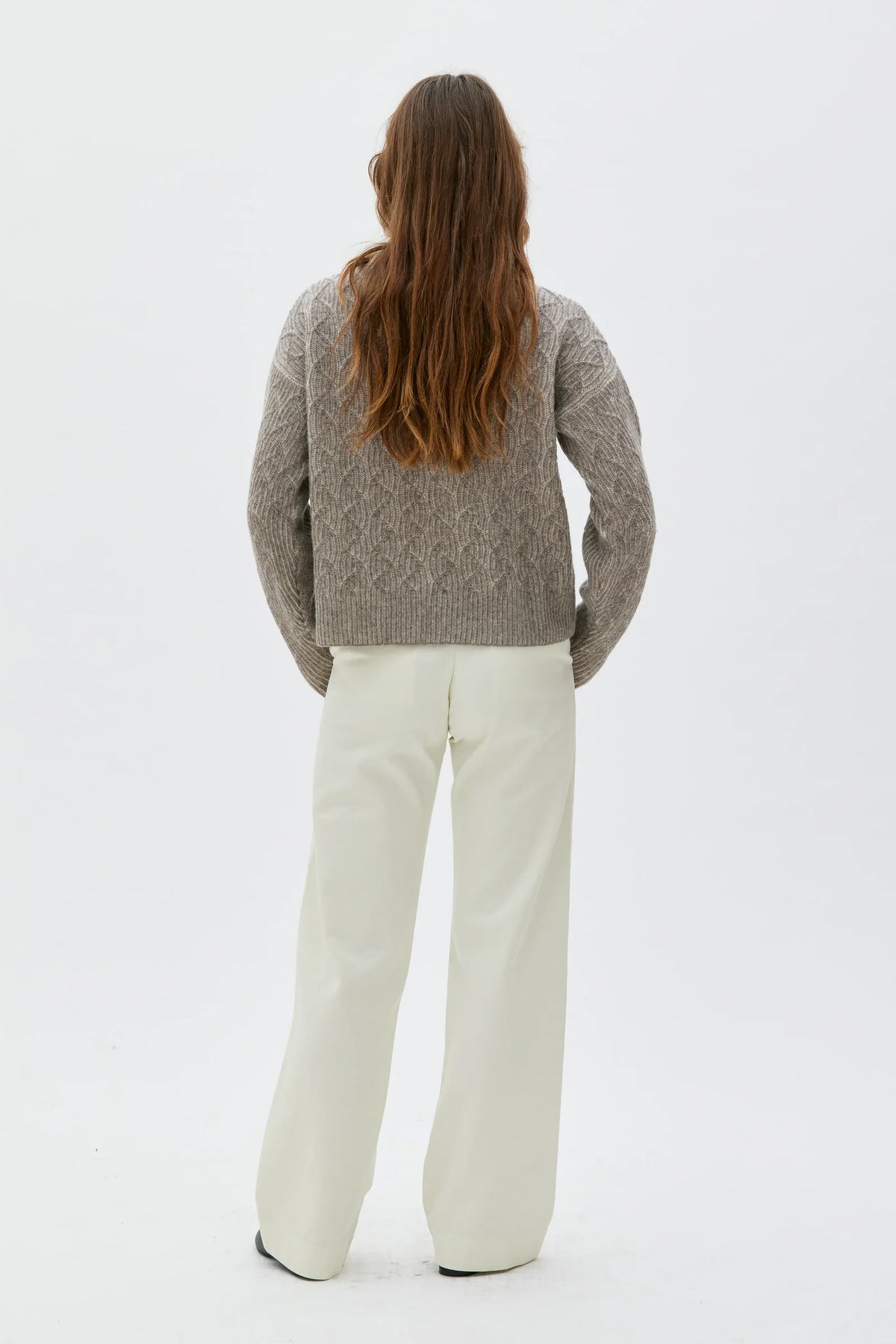 A woman viewed from behind wearing a textured taupe cable-knit sweater and off-white wide-leg trousers, standing against a seamless white studio background.