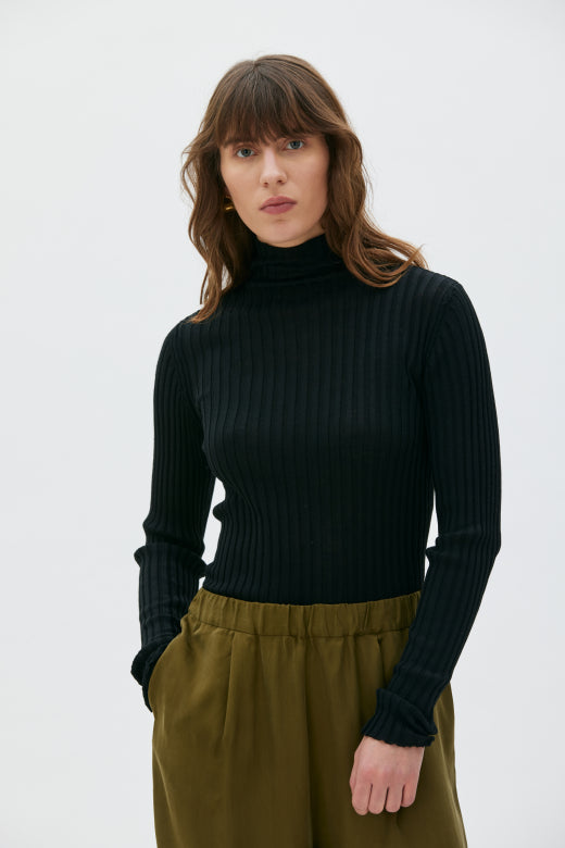 Woman wearing a black ribbed turtleneck sweater and green pants on a white background