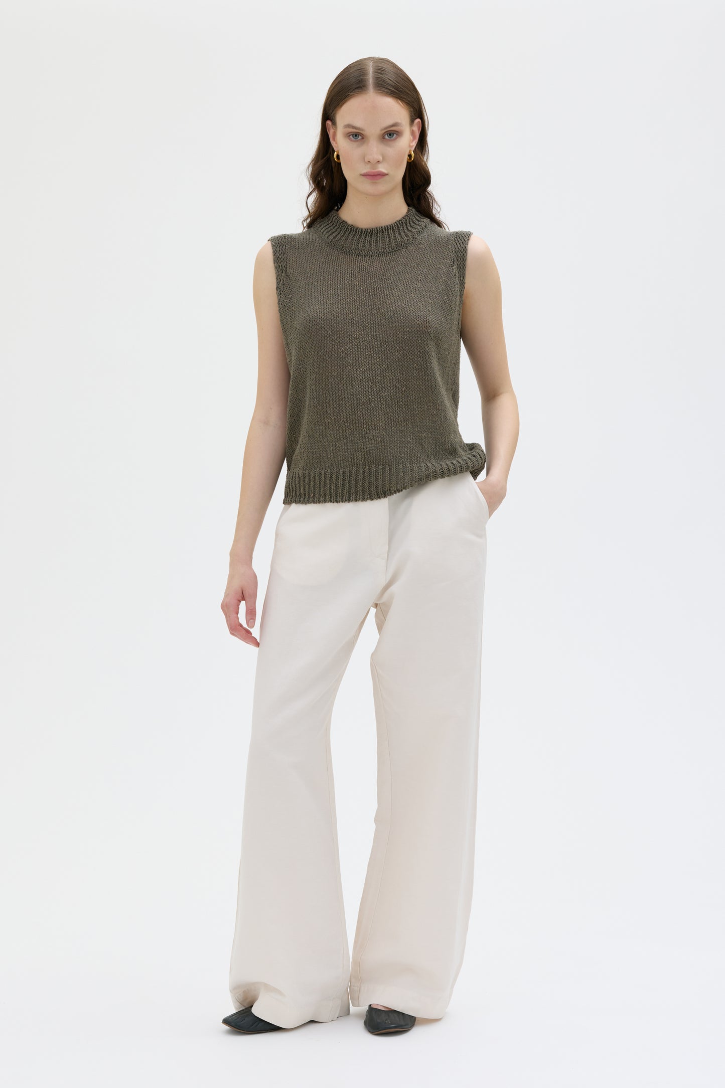Woman wearing a green sleeveless sweater and white pants on a white background