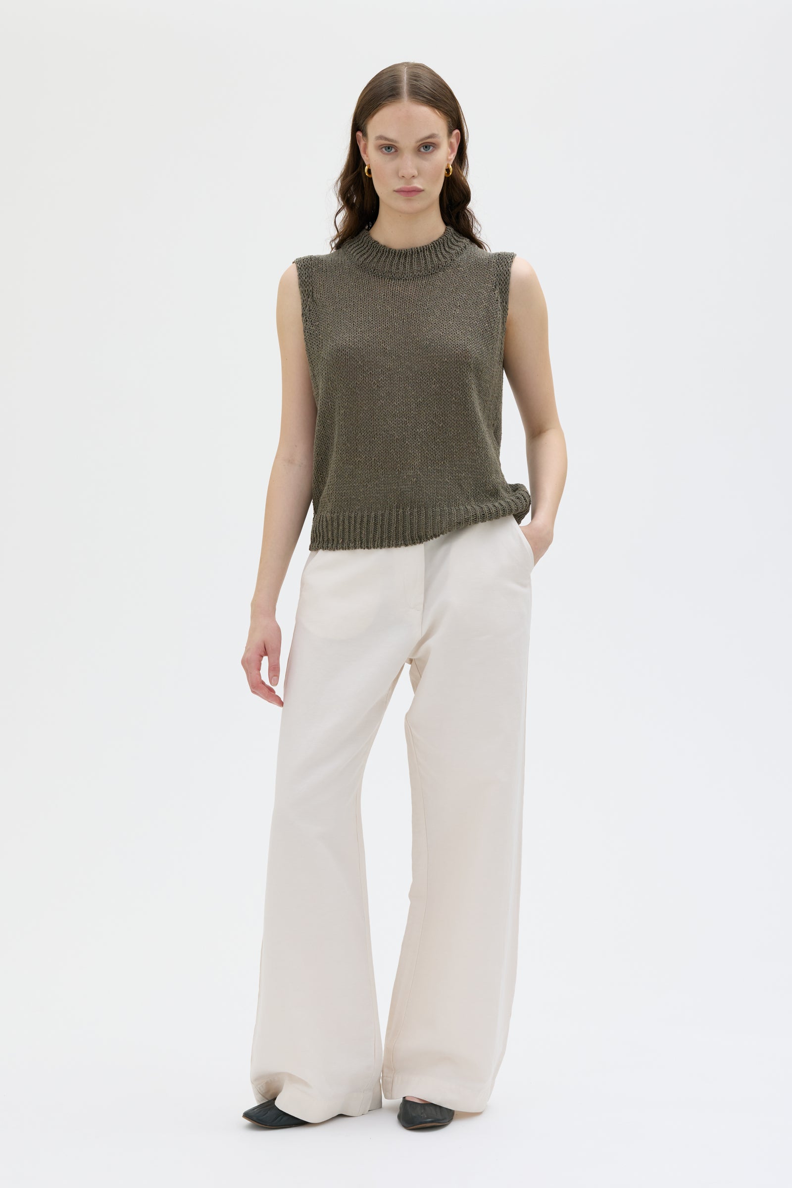 Woman wearing a green sleeveless sweater and white pants on a white background
