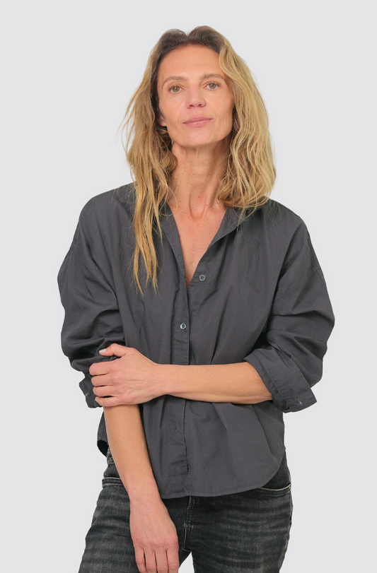 A woman with blonde hair wearing a dark gray button-down shirt and dark jeans, standing against a light gray background.