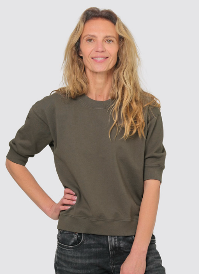 A woman with blonde hair wearing an olive green crewneck sweatshirt and dark jeans, standing against a light gray background.