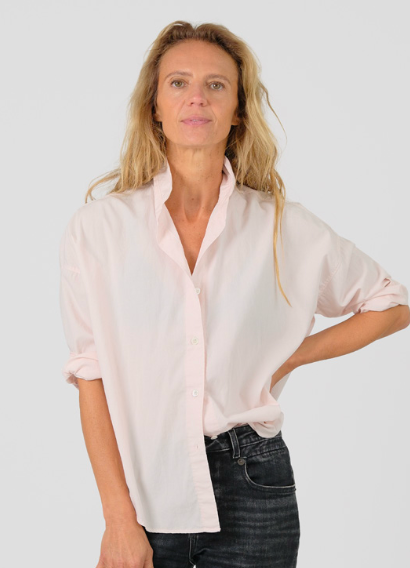 A woman modeling a light pink button-down shirt with rolled-up sleeves and dark jeans, photographed against a plain light gray studio background.