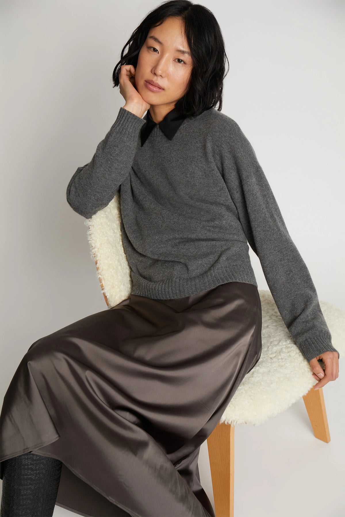 A model posing on a chair and wearing a long metallic grey skirt and a grey long-sleeve sweater.