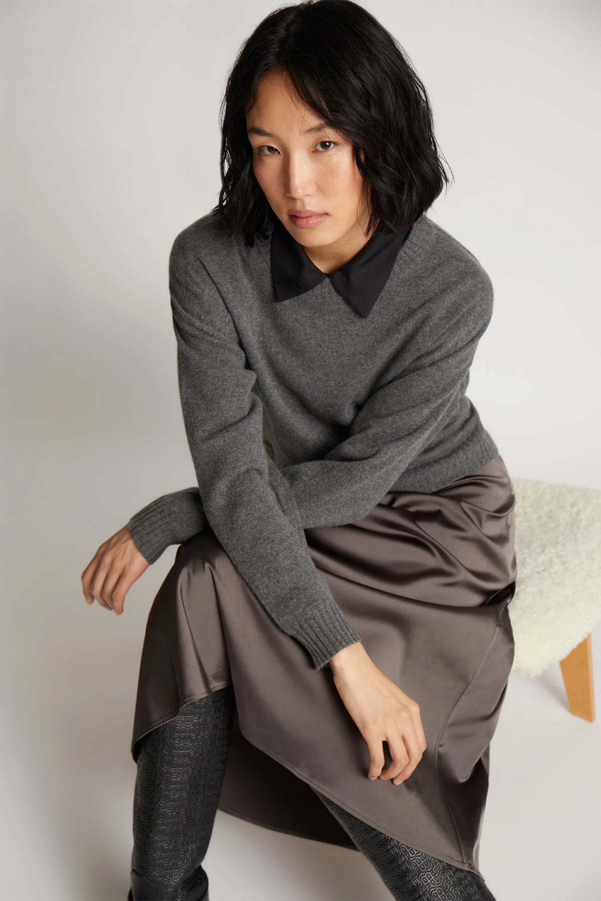 A model posing on a chair and wearing a long metallic grey skirt and a grey long-sleeve sweater.