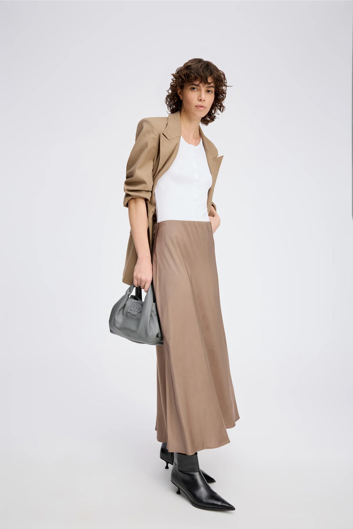 A posing model wearing a long camel color skirt, white top, and camel jacket.
