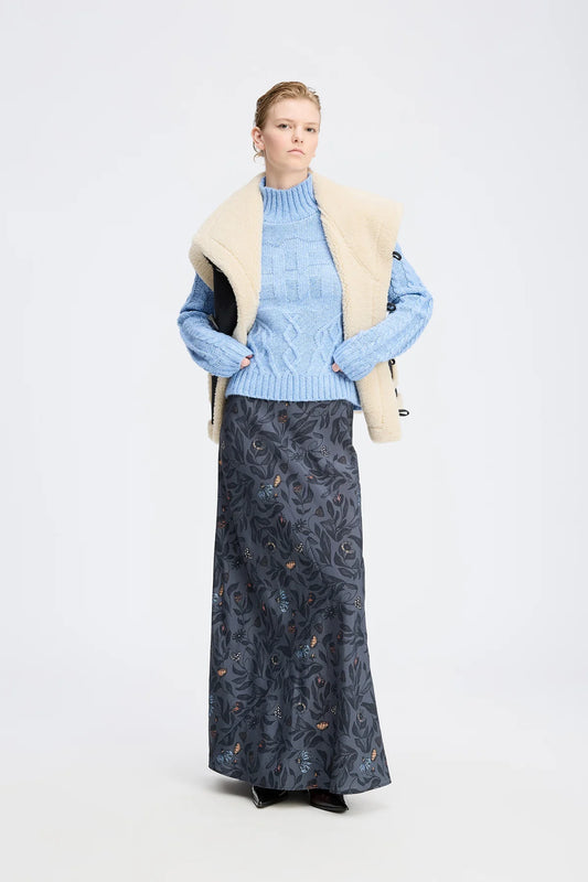 Woman wearing a light blue sweater with a cream vest over it, paired with a dark patterned skirt on a white background.