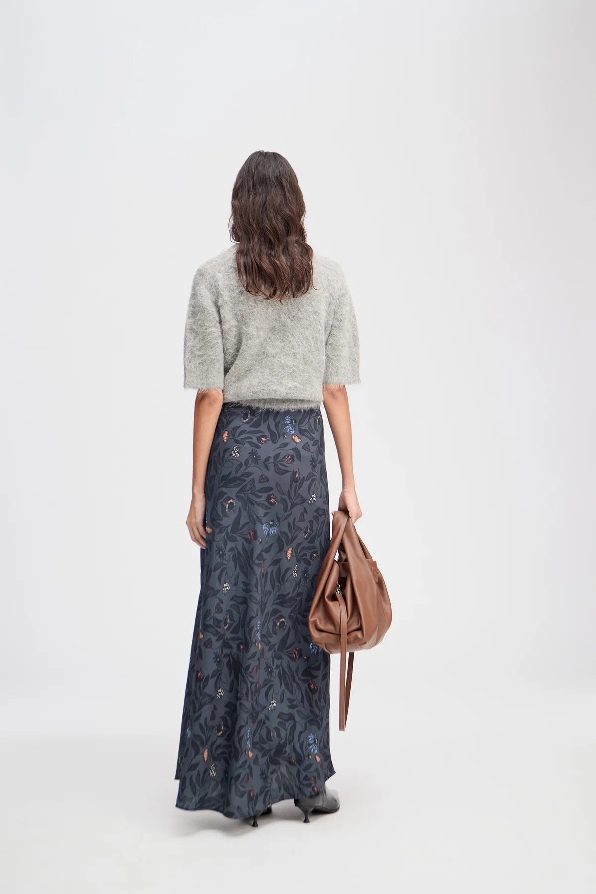 Woman wearing a gray sweater and long dark patterned skirt, holding a brown bag on a white background