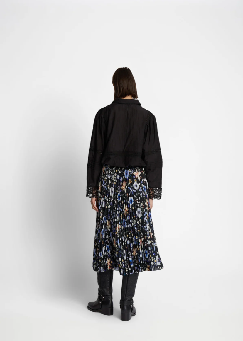 Reverse view of model wearing a pleated floral print skirt with black background and blue, white and peach colored flowers. The skirt has an elastic waistband. Skirt is work with a long sleeved black shirt.