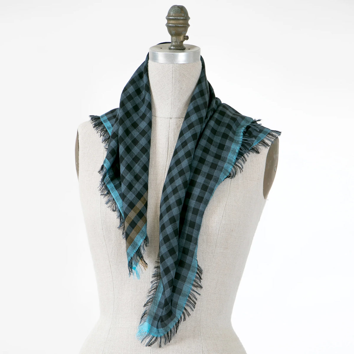 A dark green and black checkered scarf with turquoise blue edges and fringe, draped on a cream-colored fabric mannequin torso against a white background.