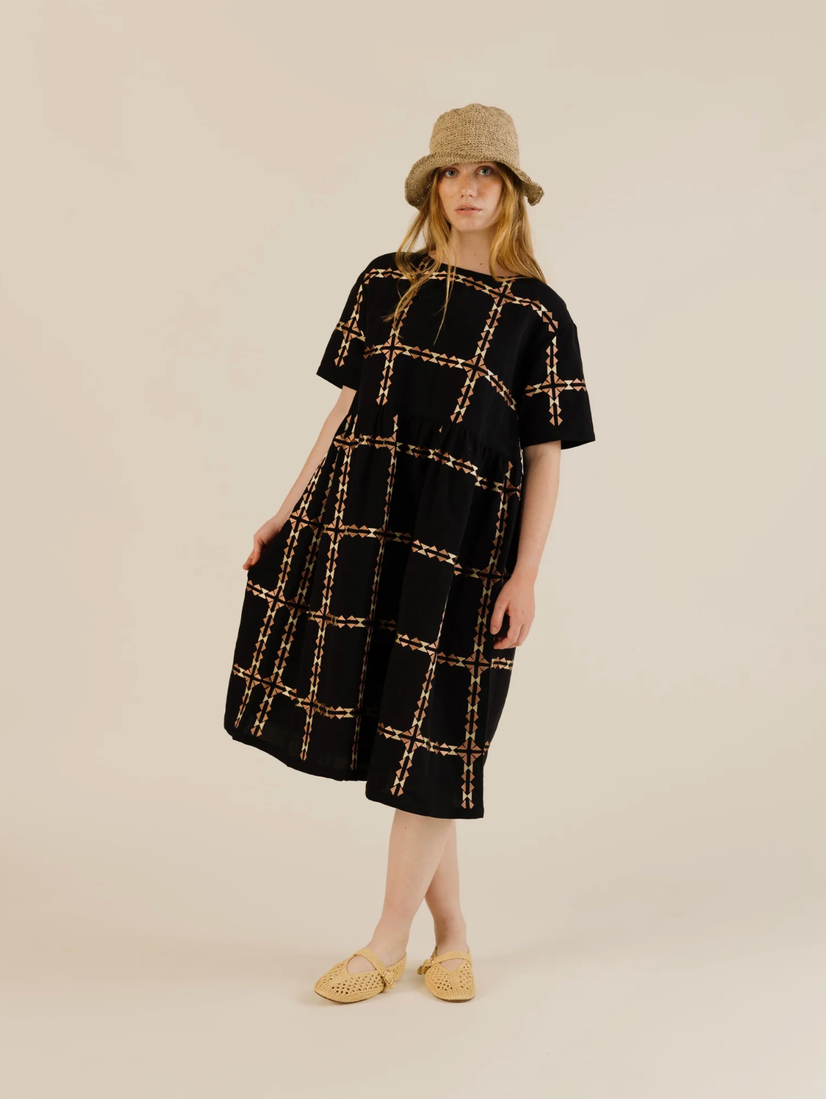 Woman wearing a black dress with gold grid pattern and a beige hat on a beige background