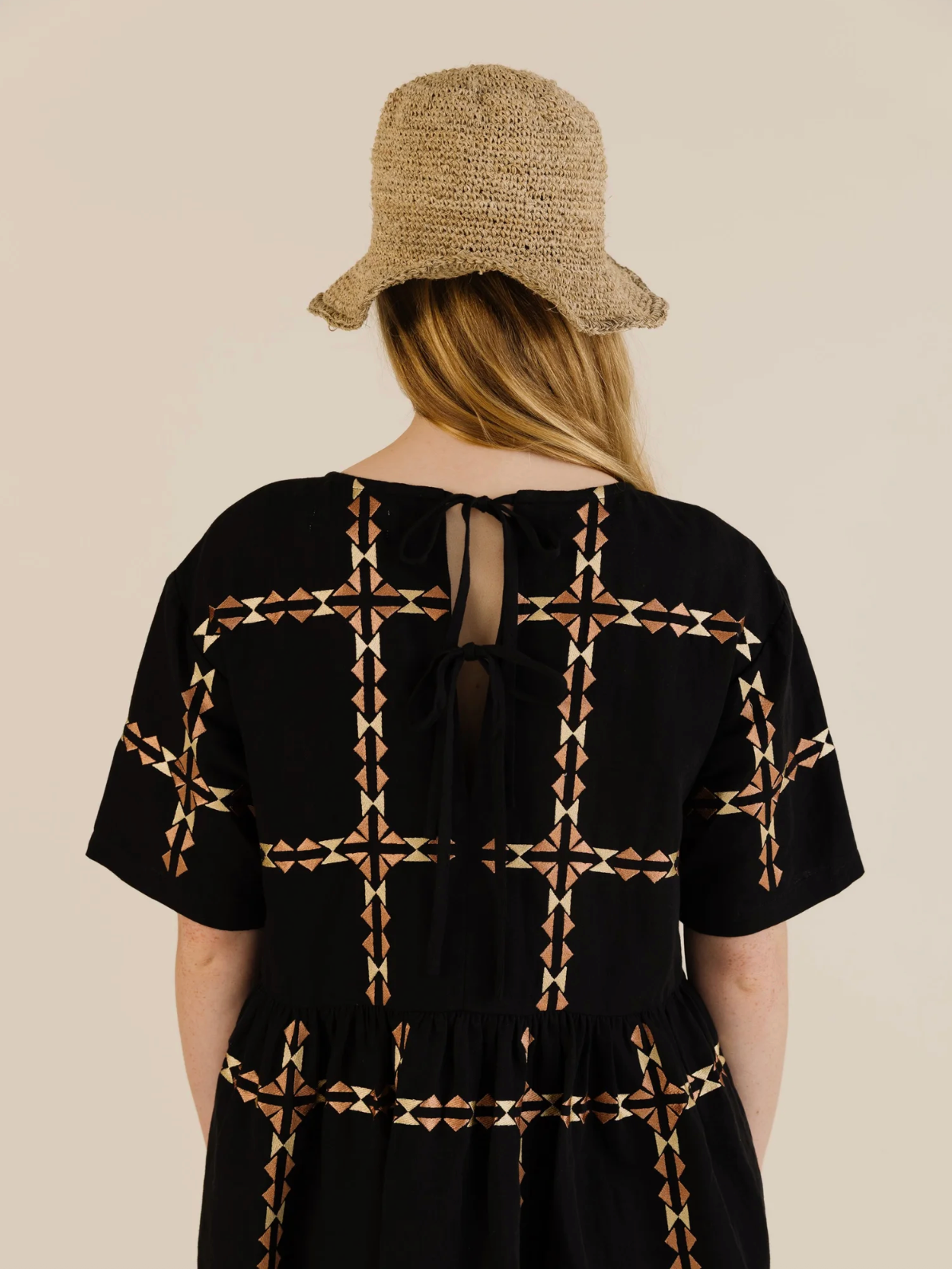 Person wearing a black dress with geometric pattern and a straw hat on a beige background