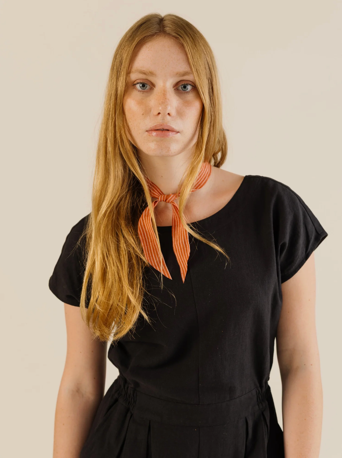 Woman wearing a black outfit with an orange scarf around her neck against a beige background