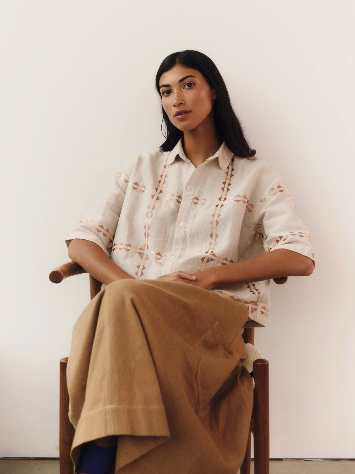 Woman sitting on a chair wearing a white embroidered shirt and brown pants against a plain background