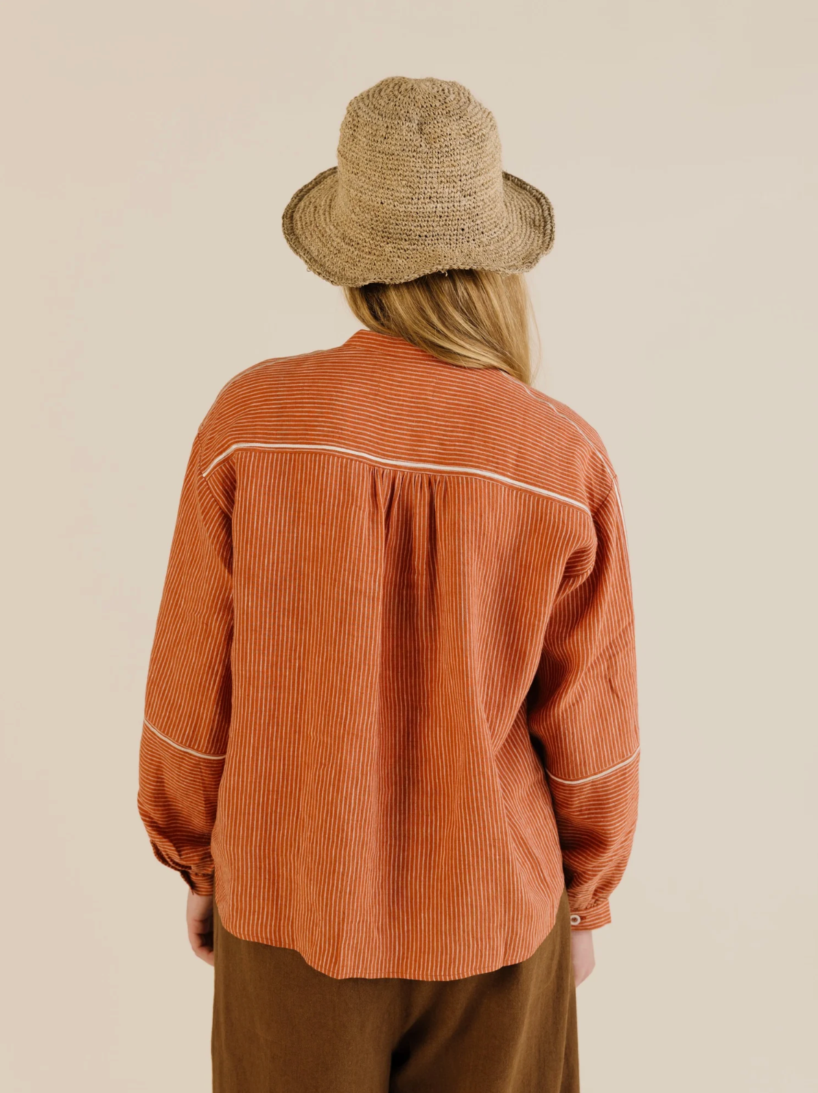 Person wearing a rust-colored corduroy shirt and brown pants with a straw hat on a beige background
