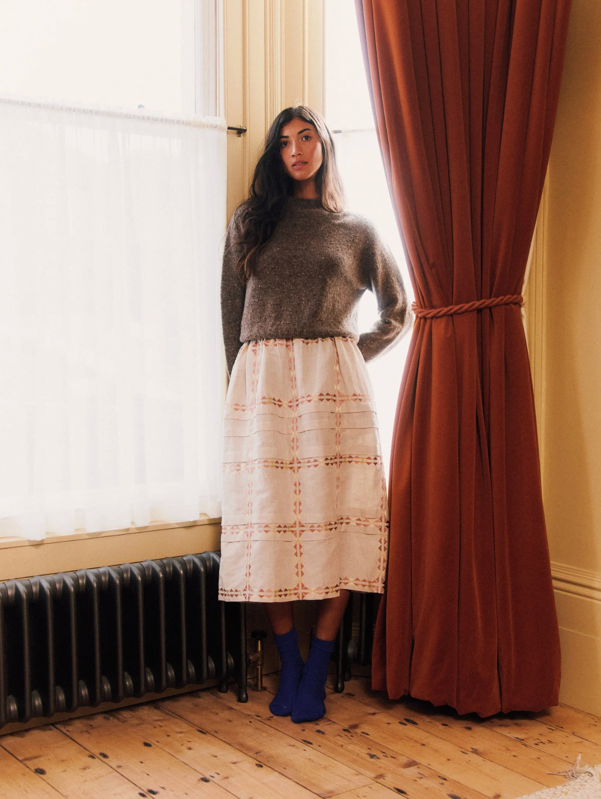 Woman standing by a window with a curtain, wearing a sweater and skirt.
