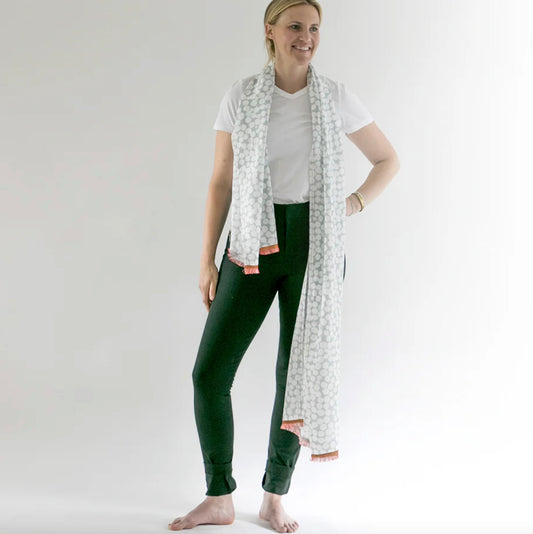 Woman wearing a long white scarf with a pattern, green pants, and a white shirt on a plain background