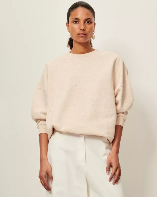 Woman wearing a beige sweater and white pants against a plain background