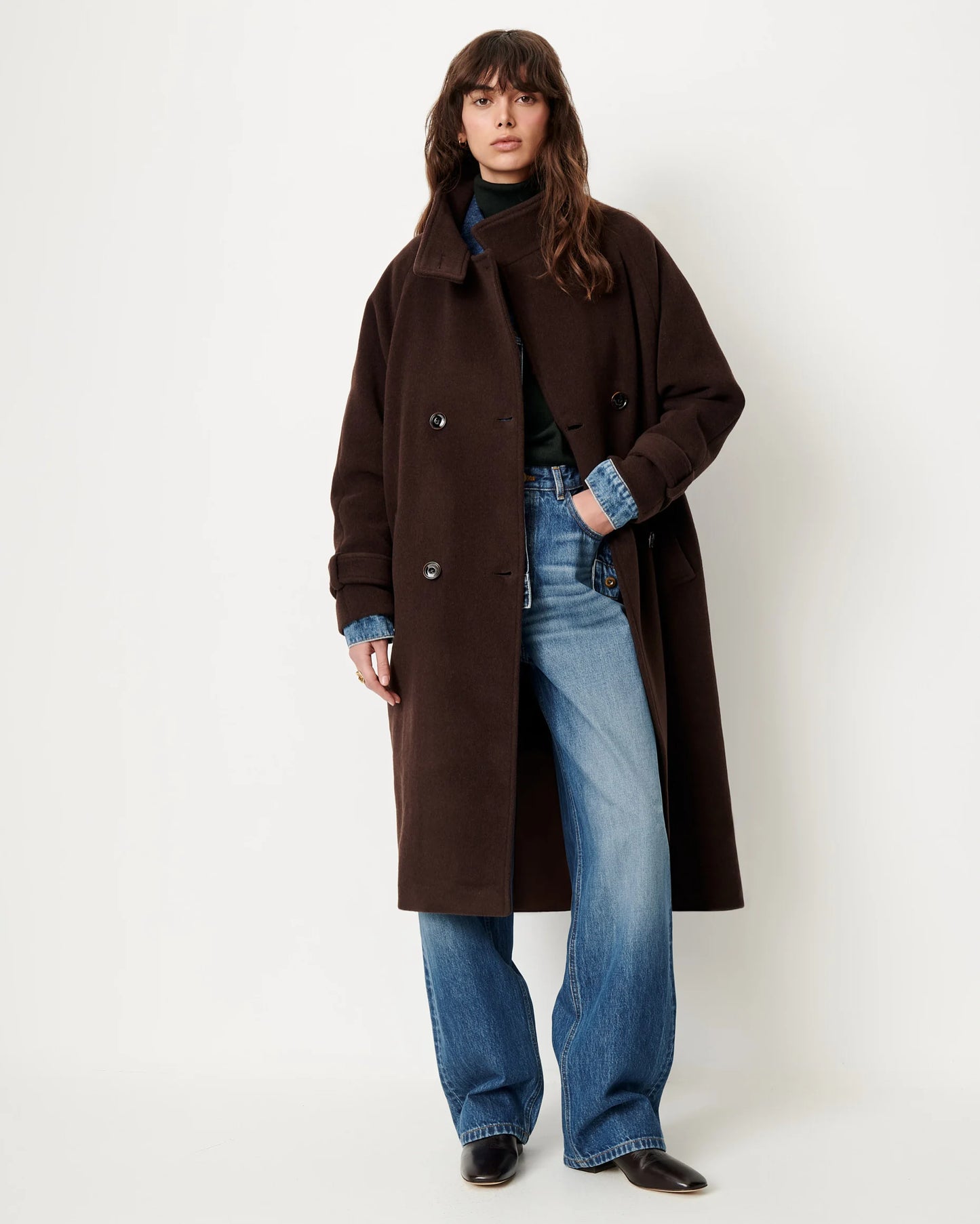 A model wearing a long, dark brown double-breasted coat over a dark green turtleneck sweater, blue jeans, and black shoes, standing against a plain off-white studio background.